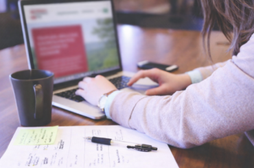 Personne travaillant sur un ordinateur portable à une table, avec un carnet de notes, un stylo et une tasse de café à côté.