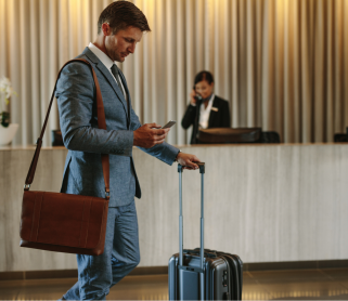 Homme en costume avec une valise consultant son téléphone dans le hall d’un hôtel, devant une réception.